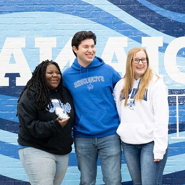 Three students standing in front of a blue-and-white painted Indiana State mural. On the far left is a white female student with straight blonde hair. She wears a white Indiana State sweatshirt, black glasses, and blue jeans. In the middle is a white male student with black hair. He wears a blue Sycamores sweatshirt and blue jeans. On the right is a Black female student with black dreadlocks. She wears a black Sycamores sweatshirt and blue jeans.